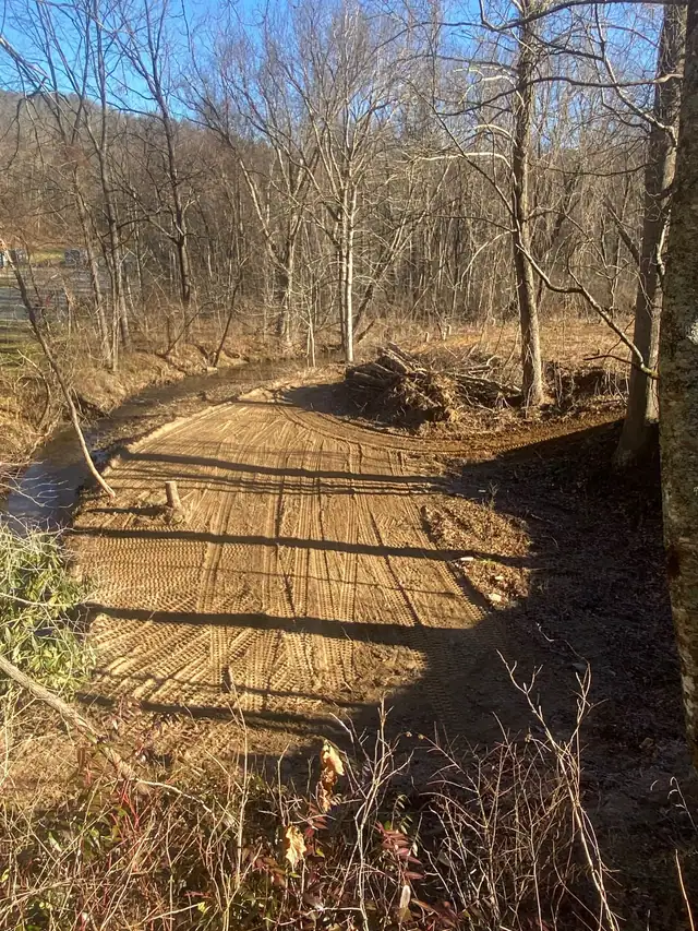Cleared, graded ground beside a creek with a brush pile at the tree line